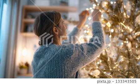 Adult woman decorates a Christmas tree with string lights, creating a cozy festive atmosphere in a beautifully arranged living space 133343505