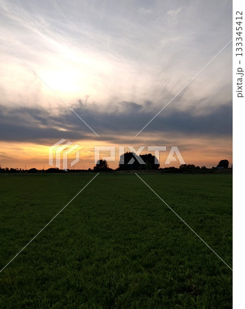 Dramatic Sunset Over Green Field with Silhouetted Trees. Stunning sunset with warm orange and pink sky over vast green field. Dark silhouettes of trees and dramatic clouds on horizon. Dramatic Sunset Over Green Field with Silhouetted Trees. Stunning sunset with warm orange and pink sky over vast green field. Dark silhouettes of trees and dramatic clouds on horizon. 133345412