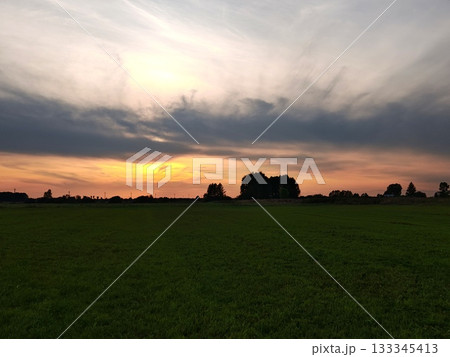 Dramatic Sunset Over Green Field with Silhouetted Trees. Stunning sunset with warm orange and pink sky over vast green field. Dark silhouettes of trees and dramatic clouds on horizon. 133345413