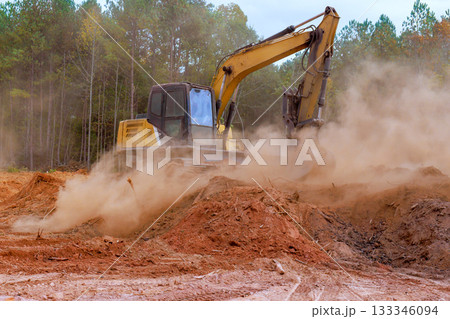 Excavator machinery digs into earth at construction site, sending dust into air among trees. 133346094