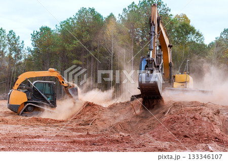 Heavy excavator machinery skid steer is working to clear dirt debris in wooded location, creating dust clouds in process. 133346107