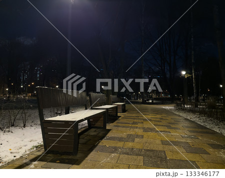 The first snow in the evening park. Snow covered wooden benches illuminated by streetlights. Park landscape with benches in snow and frozen trees. The first evening in the park after snow fell The first snow in the evening park. Snow covered wooden benches illuminated by streetlights. Park landscape with benches in snow and frozen trees. The first evening in the park after snow fell 133346177