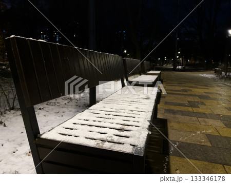 The seat of wooden bench is covered with the first snow of the year. Evening park with streetlights shining on the frozen bench, covered in ice and snow. It's impossible to sit on the cold bench The seat of wooden bench is covered with the first snow of the year. Evening park with streetlights shining on the frozen bench, covered in ice and snow. It's impossible to sit on the cold bench 133346178