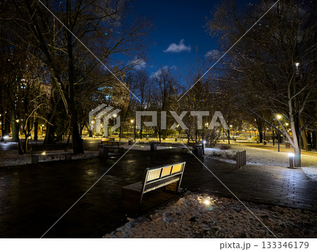 Evening park with walking areas and wooden benches covered in snow. First day of winter with fresh snow on the park grounds. Evening in a gloomy park in cold, snowy weather Evening park with walking areas and wooden benches covered in snow. First day of winter with fresh snow on the park grounds. Evening in a gloomy park in cold, snowy weather 133346179