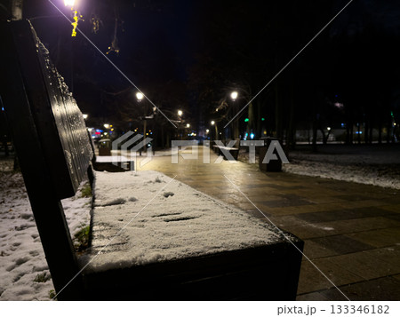 Snow covered bench in park at night. Cold, snowed park next to pedestrian area. Pedestrian walkway in the park along snow covered grass and leafless trees on the first cold days of winter 133346182