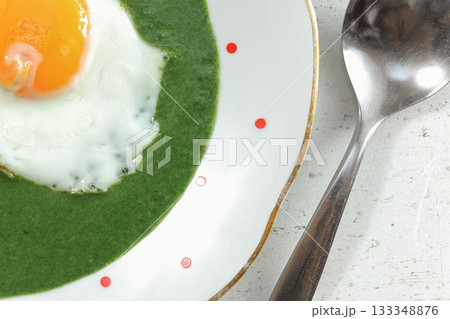 Sunny side up made egg in spinach on white plate with small red dots, steel spoon near, closeup overhead photo 133348876