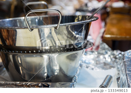 Shiny stainless steel mixing bowl and colander at a food stall, surrounded by foil, utensils, and pastry trays. Vendors prepare warm seasonal treats in a busy, atmosphere of  festive market 133349013