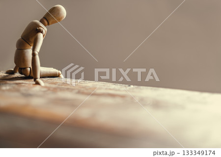 Wooden mannequin is sitting on a weathered surface with head bowed, depicting feelings of isolation, loneliness, and depression against a minimalist background. Wooden mannequin is sitting on a weathered surface with head bowed, depicting feelings of isolation, loneliness, and depression against a minimalist background. 133349174