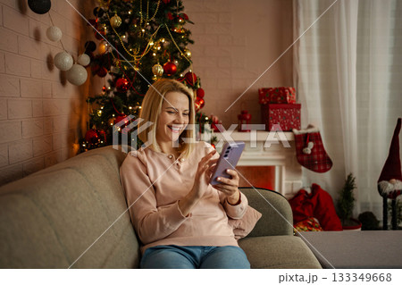 Joyful holiday moments spent on a cozy couch as a woman connects with loved ones via smartphone by a beautifully decorated Christmas tree Joyful holiday moments spent on a cozy couch as a woman connects with loved ones via smartphone by a beautifully decorated Christmas tree 133349668