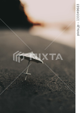 Miniature beach scene with a small umbrella on a sand at sunset. A small, white beach umbrella stands on dark sand, with a blurred sunset in the background, creating a peaceful and serene sunset scene 133349683