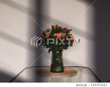 A minimalist still life of a bouquet of colorful flowers in a green glass vase standing on a woven placemat. Window shadows. Cozy home interior.. A minimalist still life of a bouquet of colorful flowers in a green glass vase standing on a woven placemat. Window shadows. Cozy home interior.. 133351012
