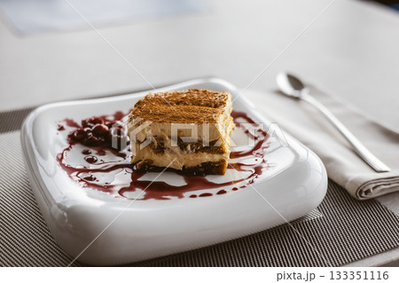 Delicious tiramisu dessert with cherries served on a white plate with a berry sauce. A square of tiramisu dusted with cocoa powder 133351116
