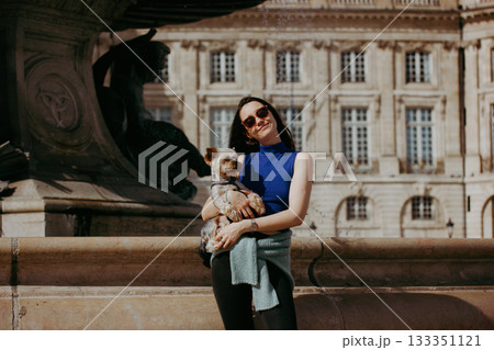 Young brunette woman holding a small dog Yorkshire terrier in front of a fountain. A girl with sunglasses holds a doggy in her arms, posing on a city street on a sunny day. A trip with a lapdog, pet. 133351121