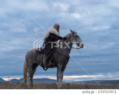 A Viking-like chieftain with a white beard and fur cloak sits contemplatively on a dappled grey horse, gently touching its head, under a dramatic cloudy sky over mountains. 133352913