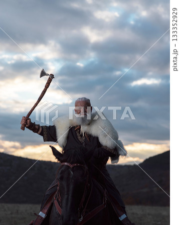 A fierce Viking warrior on horseback, wielding an axe, stands against a dramatic sunset sky with clouds and mountains, embodying strength, exploration, and ancient legend. 133352929