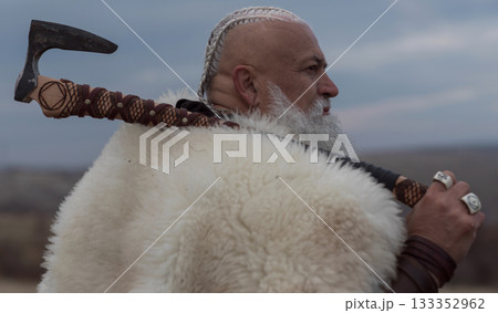 Viking warrior with axe standing on mountain cliff, wearing fur cloak and leather armor, overlooking dramatic landscape. Historical Nordic fighter 133352962