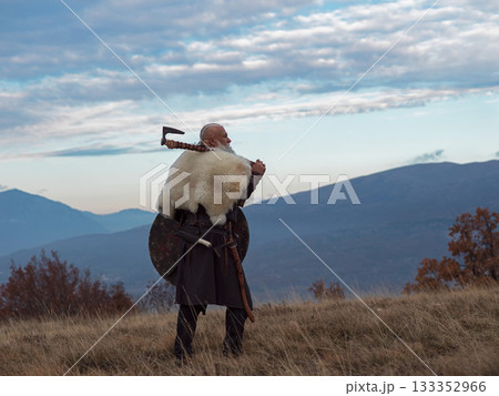 Viking warrior with axe standing on mountain cliff, wearing fur cloak and leather armor, overlooking dramatic landscape. Historical Nordic fighter 133352966