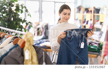 Happy pretty young girl client carefully choosing stylish jeans trousers in clothing store. Winter sales concept 133352987