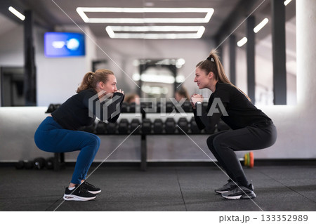 Two women perform squats together in a modern gym during a fitness session focused on strength training and camaraderie Two women perform squats together in a modern gym during a fitness session focused on strength training and camaraderie 133352989