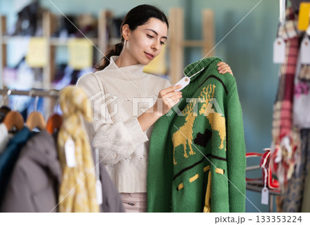Interested young girl buyer standing in clothing market and carefully examines selection of sweaters on sale of winter clothing collection Interested young girl buyer standing in clothing market and carefully examines selection of sweaters on sale of winter clothing collection 133353224