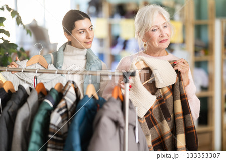 Family shopping concept. Two happy women choosing coat or jacket for cold season in clothing shop with large assortment Family shopping concept. Two happy women choosing coat or jacket for cold season in clothing shop with large assortment 133353307