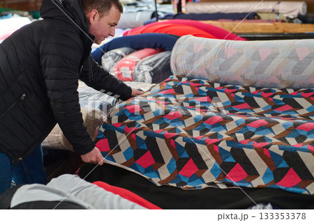 Designer selecting colorful patterned fabric rolls in a textile warehouse, choosing materials for sewing, fabric store, textile industry, material selection, indoor shot. 133353378
