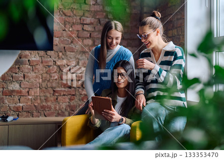 Three cheerful young women sitting in a cozy modern interior, smiling and looking at a tablet screen together, representing friendship, technology, and digital lifestyle. Three cheerful young women sitting in a cozy modern interior, smiling and looking at a tablet screen together, representing friendship, technology, and digital lifestyle. 133353470