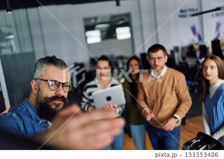 Group of young professionals gathered in a modern office for a business presentation. A man with a beard explains data charts on a large screen while the team listens attentively. Concept of teamwork Group of young professionals gathered in a modern office for a business presentation. A man with a beard explains data charts on a large screen while the team listens attentively. Concept of teamwork 133353486