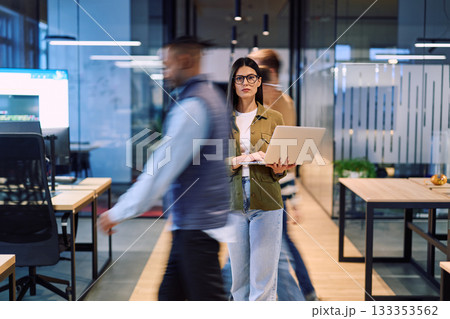 Business woman holding laptop in the middle of a dynamic modern office while colleagues move around, symbolizing focus, leadership, and productivity amid a fast-paced work environment. 133353562