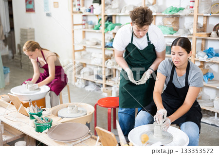 Young woman teacher helps teenagers boy and girl to make product on potter's wheel 133353572