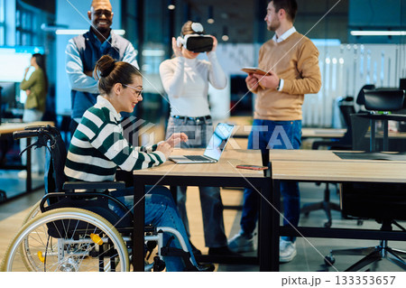 Diverse business team collaborating in a modern office environment. A woman in a wheelchair works on a laptop while colleagues explore virtual reality and digital tools, promoting innovation 133353657