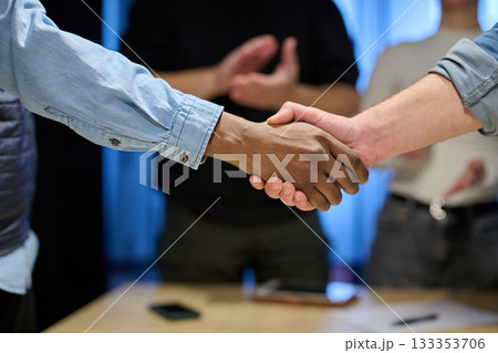 Two businessmen shaking hands after successful negotiation in a modern office, symbolizing partnership, agreement, success, and teamwork. A woman in the background applauds the deal. 133353706
