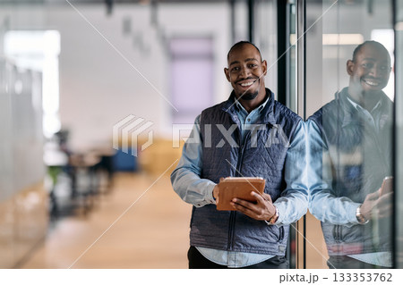 Confident african american businessman standing in a modern office hallway, smiling while using a digital tablet. Reflection in glass wall symbolizes technology, success, and innovation in business. Confident african american businessman standing in a modern office hallway, smiling while using a digital tablet. Reflection in glass wall symbolizes technology, success, and innovation in business. 133353762