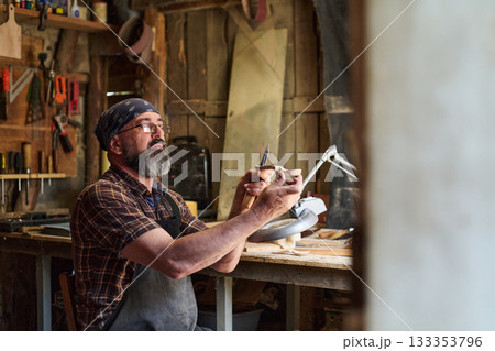 Craftsman carving wood in workshop, artisan shaping wooden object with knife, skilled woodworker creating handmade craft, hobbyist at work in rustic studio. 133353796