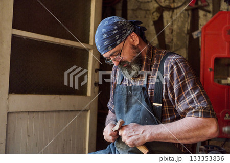 Craftsman carving wood in workshop, artisan shaping timber with knife, woodworking concept, carpentry skills, handmade craft, hobby and occupation, vintage setting, natural light. Craftsman carving wood in workshop, artisan shaping timber with knife, woodworking concept, carpentry skills, handmade craft, hobby and occupation, vintage setting, natural light. 133353836