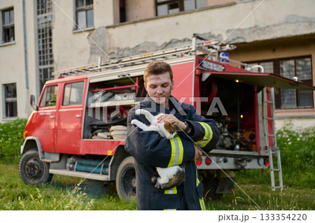 Brave firefighter holding a rescued cat in his arms near a fire truck, showing compassion and care, outdoor scene with a building in the background Brave firefighter holding a rescued cat in his arms near a fire truck, showing compassion and care, outdoor scene with a building in the background 133354220