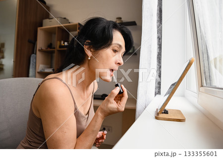 A 50 years old woman applies lip gloss at home, showing her natural face with visible wrinkles, embracing real beauty, confidence and a gentle self care moment. A 50 years old woman applies lip gloss at home, showing her natural face with visible wrinkles, embracing real beauty, confidence and a gentle self care moment. 133355601