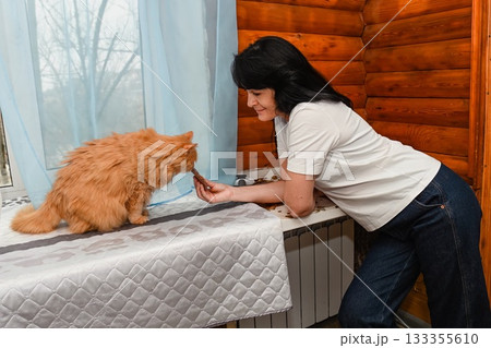 Mature 50 years old Russian woman feeds her senior 20 years old ginger cat by the window, sharing a warm and tender moment in a cozy wooden room. Mature 50 years old Russian woman feeds her senior 20 years old ginger cat by the window, sharing a warm and tender moment in a cozy wooden room. 133355610