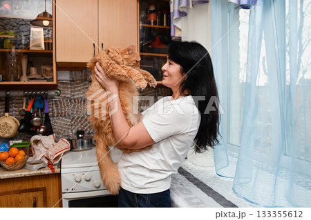 Fifty year old woman holding her twenty year old orange cat in a cozy kitchen interior, smiling by the window in natural light. Fifty year old woman holding her twenty year old orange cat in a cozy kitchen interior, smiling by the window in natural light. 133355612