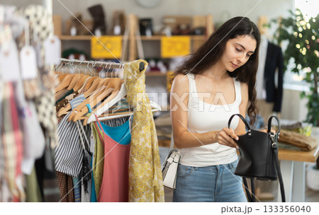 Young woman customer making choice and elect leather handbag in store Young woman customer making choice and elect leather handbag in store 133356040