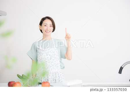 エプロン姿の主婦が笑顔で家庭料理のオススメポイントを指差し案内する明るいキッチンシーン エプロン姿の主婦が笑顔で家庭料理のオススメポイントを指差し案内する明るいキッチンシーン 133357030