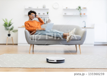 A person relaxes on a couch while a robot vacuum cleans the floor in a modern living room. 133360239