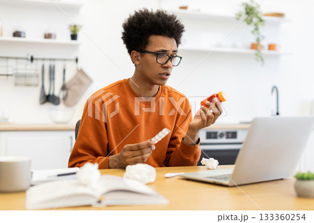 A young man, wearing glasses, looks at medication while using a laptop, possibly consulting with a doctor or researching symptoms. A young man, wearing glasses, looks at medication while using a laptop, possibly consulting with a doctor or researching symptoms. 133360254