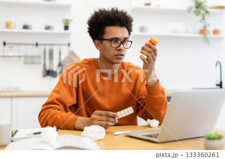 A young man with glasses looks at medicine while sitting at a table with a laptop and tissues, possibly sick. A young man with glasses looks at medicine while sitting at a table with a laptop and tissues, possibly sick. 133360261
