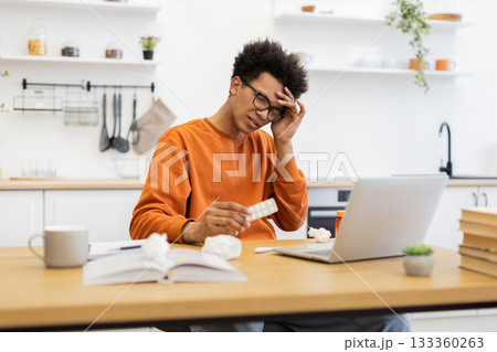 A young man, appearing unwell, holds medication while working on a laptop at home, suggesting illness and remote work. A young man, appearing unwell, holds medication while working on a laptop at home, suggesting illness and remote work. 133360263