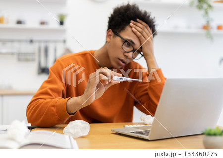 A young man, looking unwell, checks his temperature with a thermometer while working on his laptop at home. A young man, looking unwell, checks his temperature with a thermometer while working on his laptop at home. 133360275