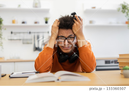 A young man with glasses is visibly frustrated, holding a game controller while sitting at a table with a book. A young man with glasses is visibly frustrated, holding a game controller while sitting at a table with a book. 133360307