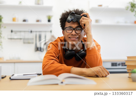 A young man wearing glasses looks frustrated while holding a game controller, with headphones around his neck and a book open in front of him. 133360317