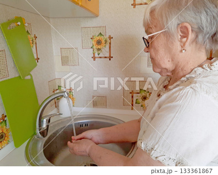 An elderly woman wearing glasses washes her hands or an object in a metal sink in the kitchen. An elderly woman wearing glasses washes her hands or an object in a metal sink in the kitchen. 133361867