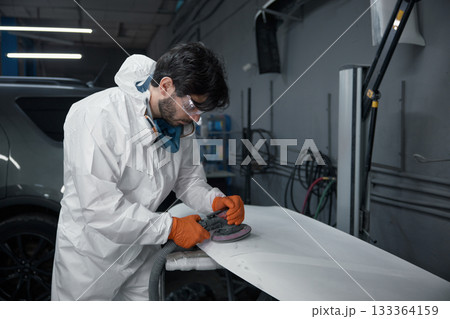 An auto body technician in a protective suit meticulously sands a car hood at a repair shop An auto body technician in a protective suit meticulously sands a car hood at a repair shop 133364159